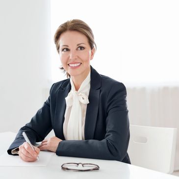 Professional woman in a suit smiling while writing at a desk with glasses in front.