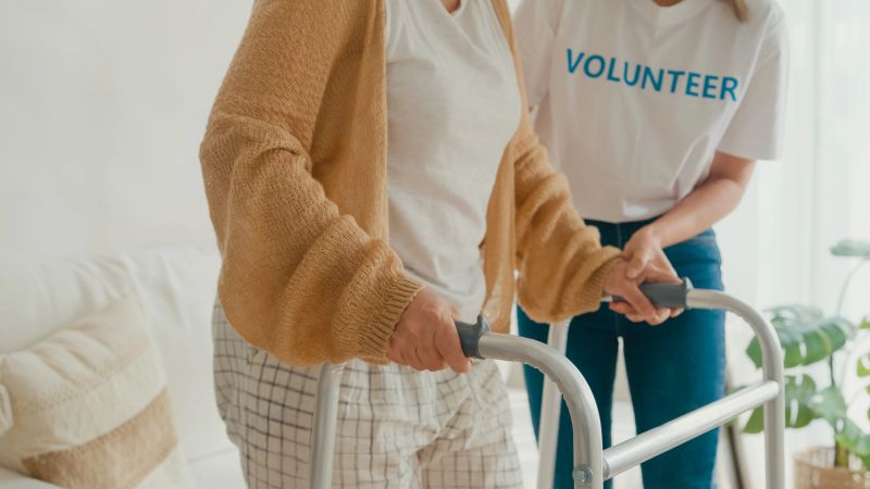 Closeup of young Asian female volunteer teach old woman to walk use walker take care hold hand elderly patient in living room at home. Volunteer health care concept.
