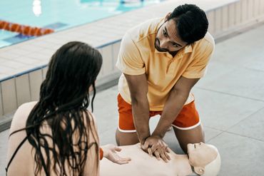 Man performing CPR on a training dummy by a poolside with a woman watching.
