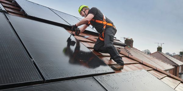 Worker installing solar panels on a sloped roof with safety gear.