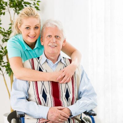 Smiling woman embracing elderly man in wheelchair at home.