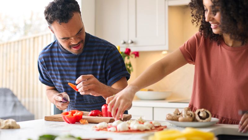 Couple At Home With Man With Down Syndrome And Woman Preparing Topping For Pizza In Kitchen Together