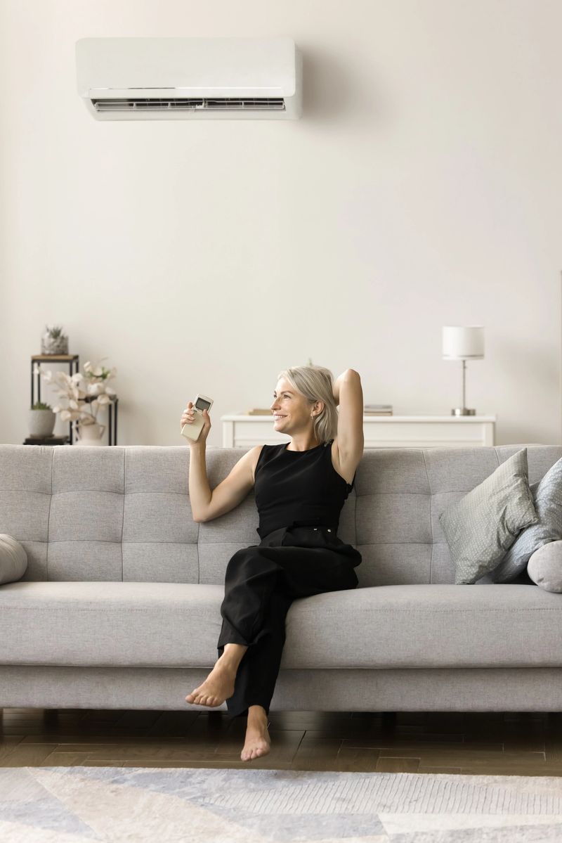 Happy relaxed middle aged woman starting air conditioner from comfortable sofa, resting on couch at home, using conditioning cooling ventilation split system with equipment on wall