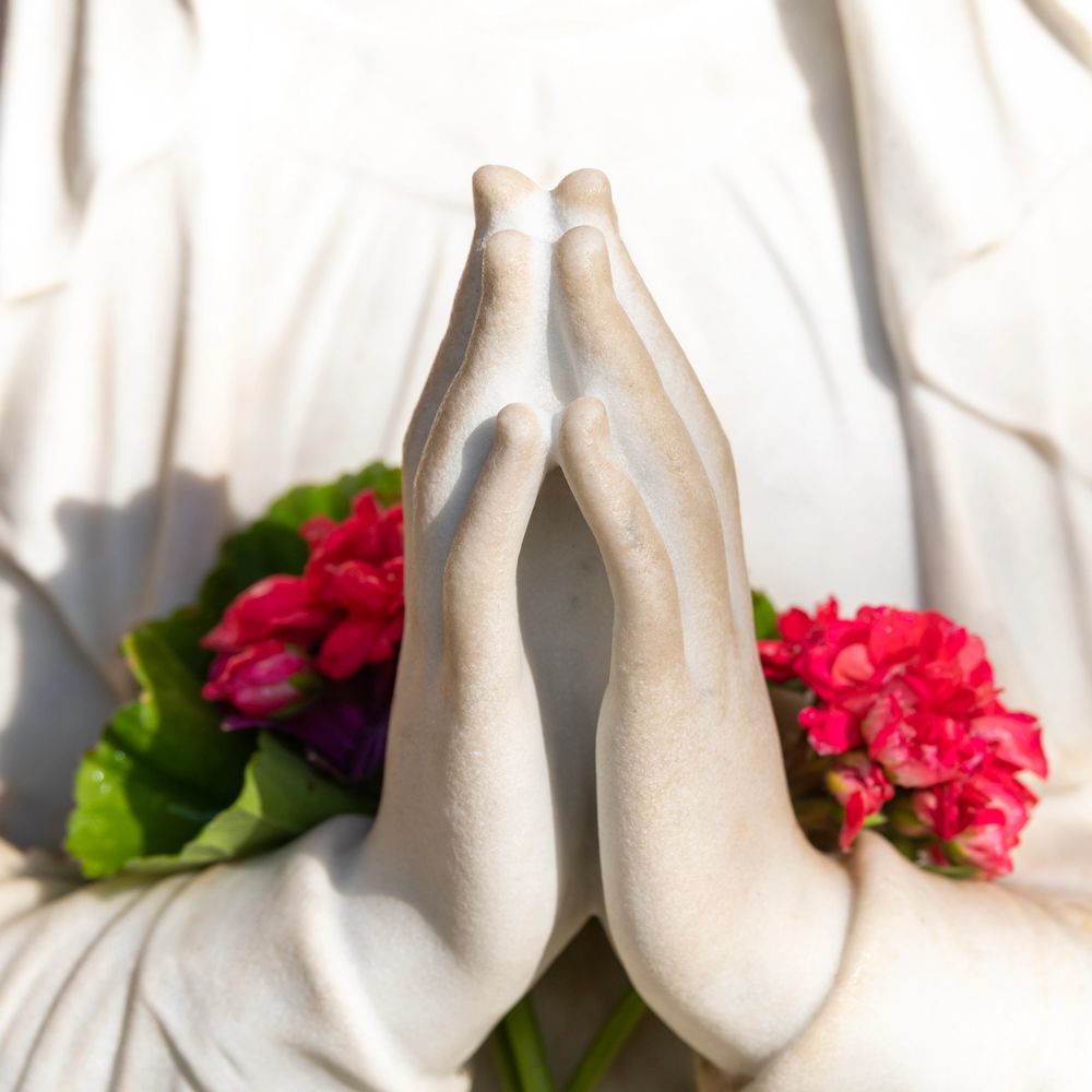 Close-up of praying hands statue with vibrant flowers in the background.