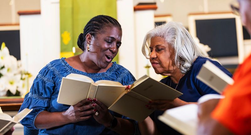 Two women in a group bible study meeting at church. A senior African-American woman, in her 70s with gray hair, is leaning over to listen to a younger woman, in her 50s.