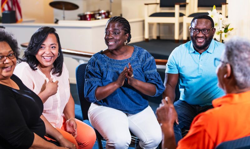 A group of black and African-American men and women sitting in a circle having a discussion. It could be a planning committee meeting, or perhaps a group therapy session. They are smiling, looking to the side. The woman in the middle is in her 50s.