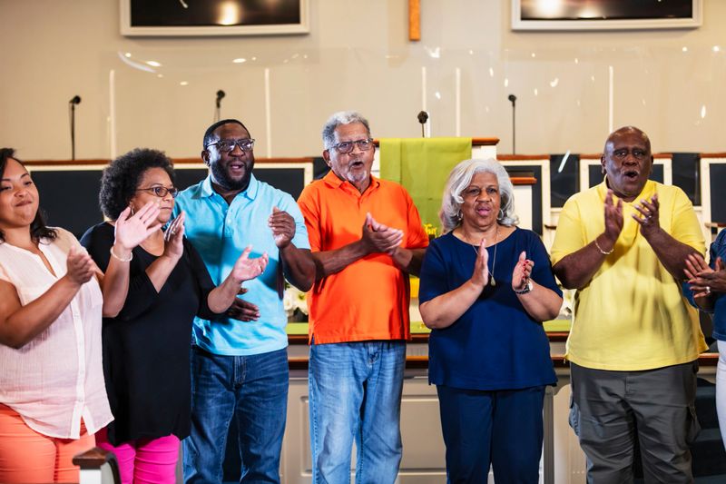 A group of six black and African-American standing together, clapping and singing. The woman on the left is in her 30s. The rest of the group are seniors.