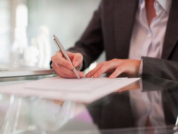 Person in business attire signing a document at a glass table.