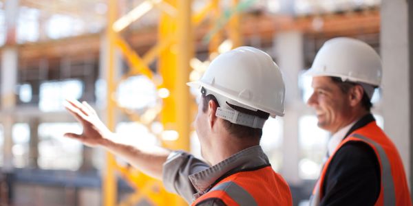 Two construction workers in helmets and vests discuss a building site.