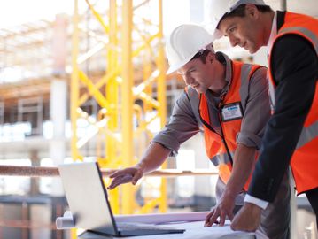 Two construction workers review plans on a laptop.