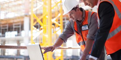Two construction engineers reviewing plans on a laptop at a building site.