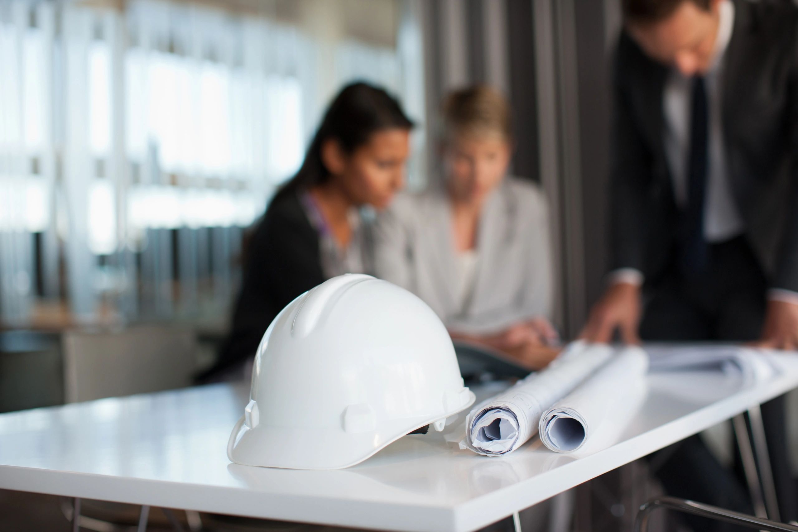 White construction helmet and blueprints on a table with professionals discussing about sustainable living spaces in background