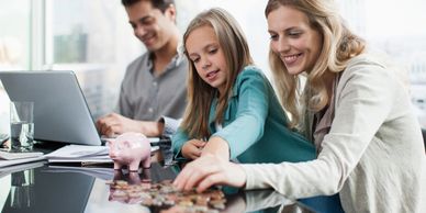 Family counting coins with a piggy bank on a table, father working on laptop.