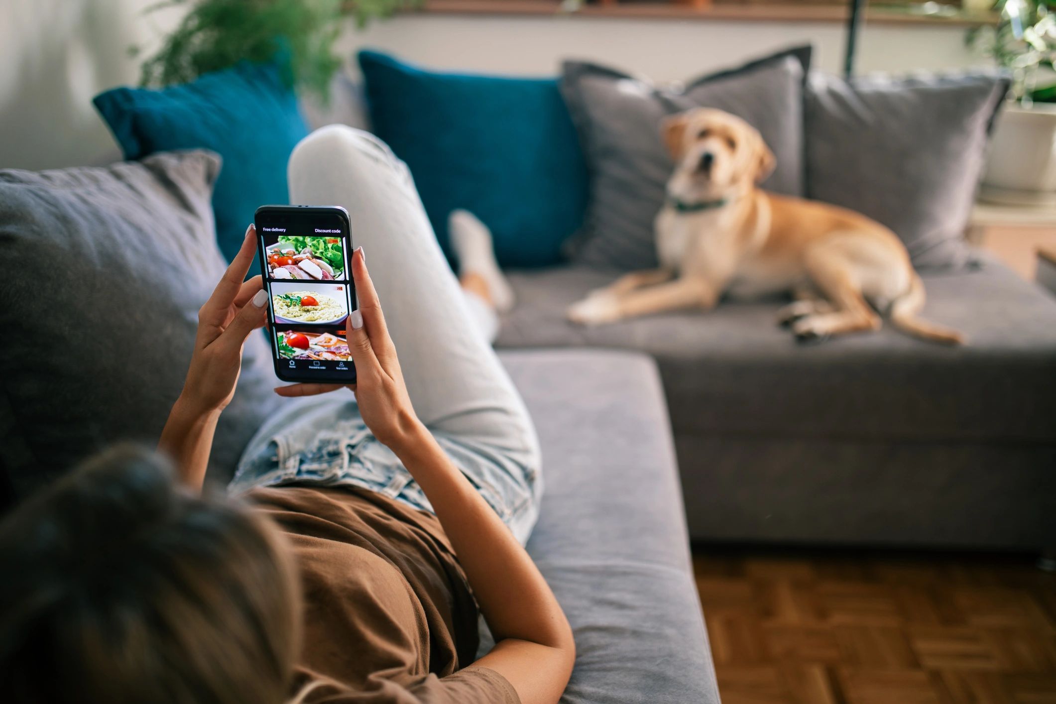 Person relaxing on couch shopping for food on phone with dog nearby.