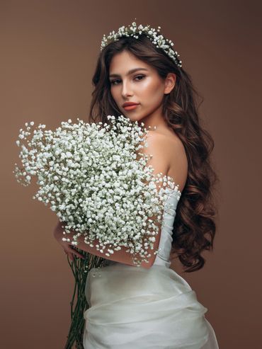 Elegant woman in white dress holding a large bouquet of white flowers against a brown backdrop.