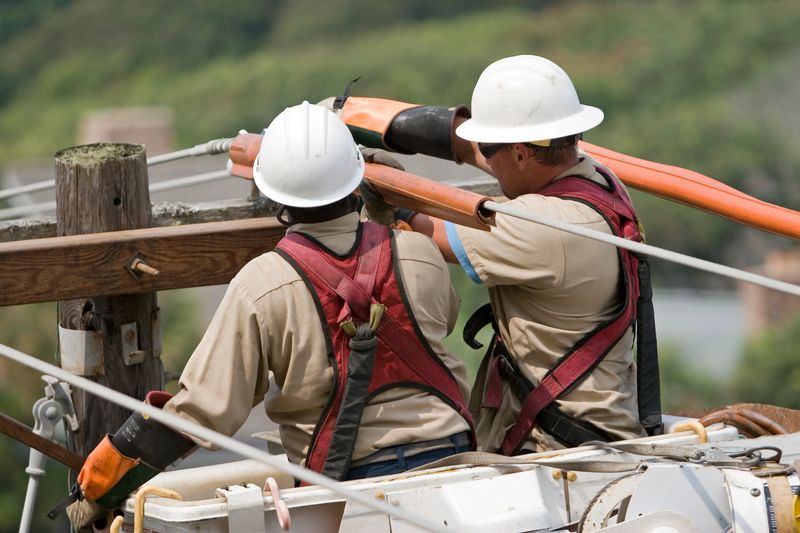 Pole-level view of electric utility workers making repairs on live transmission lines.