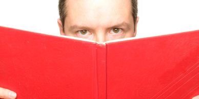 Man peeking over a large red book with focused eyes.