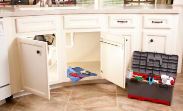 Open kitchen cabinet with plumbing tools and toolbox on floor.