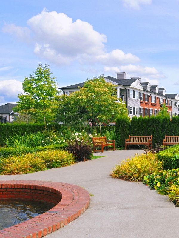 A peaceful garden path with benches and a brick water feature near residential buildings.