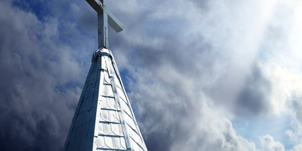 Silver church steeple with a cross under a dramatic cloudy sky.