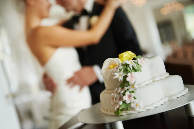 A beautiful wedding cake on a cake stand with a newlywed couple embracing in the background - focus on foreground