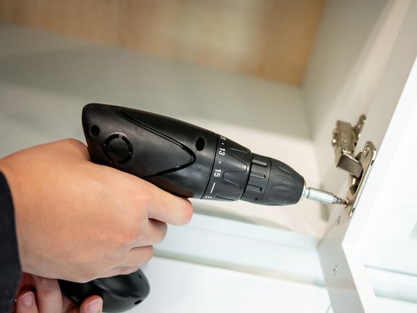 Person using a black electric drill to fix a cabinet hinge.