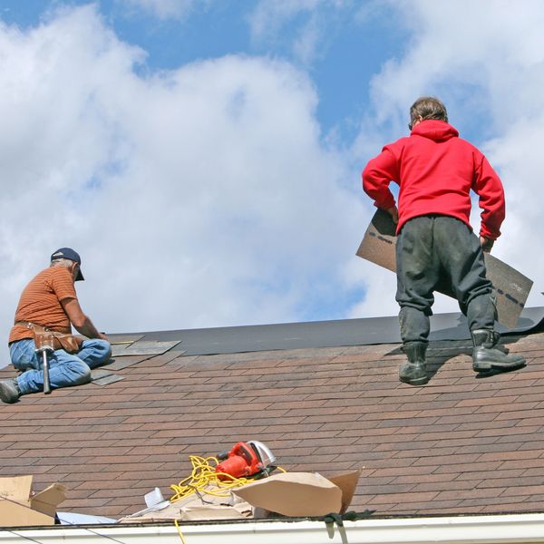 Two workers installing roofing shingles under a blue sky.
