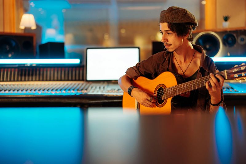Caucasian artist wearing a hat while sitting and playing guitar in the studio besides audio mixer