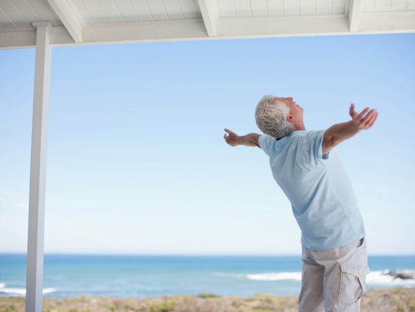 Elderly man stretching his arms wide on a porch with an ocean view.