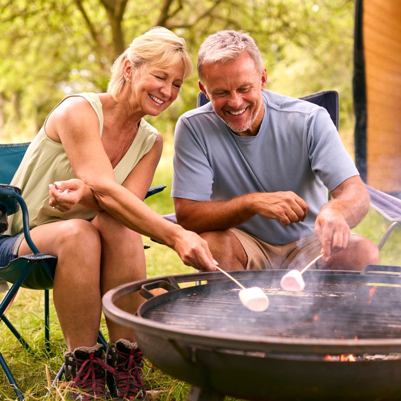 Senior Couple Camping In Countryside With RV Toasting Marshmallows Outdoors On Fire