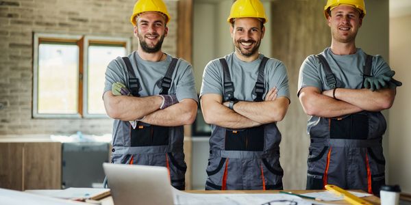 Three construction workers in helmets smiling with arms crossed indoors.