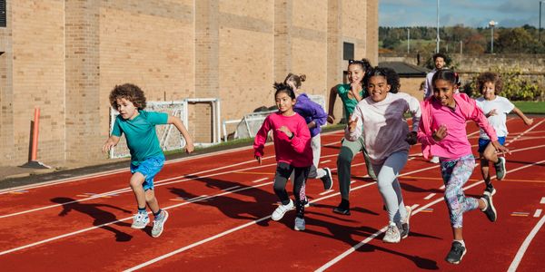 Children racing on an outdoor track with a coach in the background.