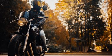 Motorcyclist in black helmet and leather jacket rides through autumn trees.