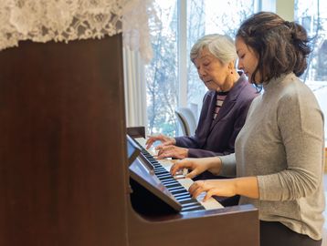 An elderly woman and a younger woman play piano together indoors.