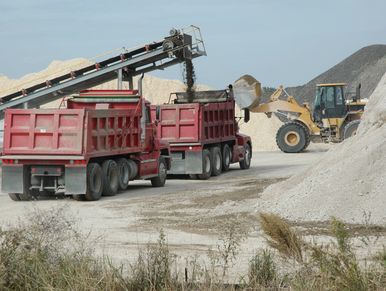 Two red dump trucks being loaded with gravel by a conveyor and a loader in a quarry.