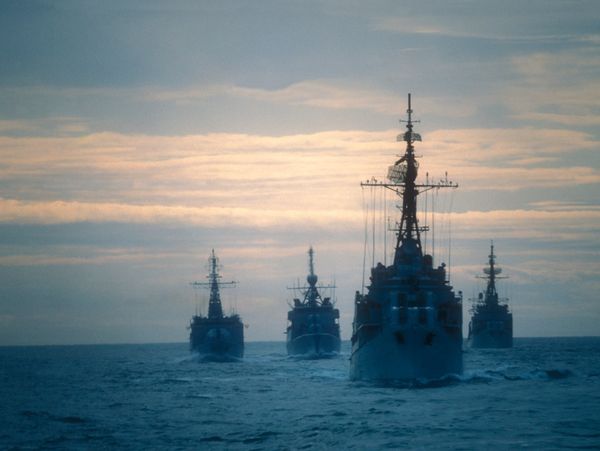 Four naval ships sailing in formation on the ocean during sunset.