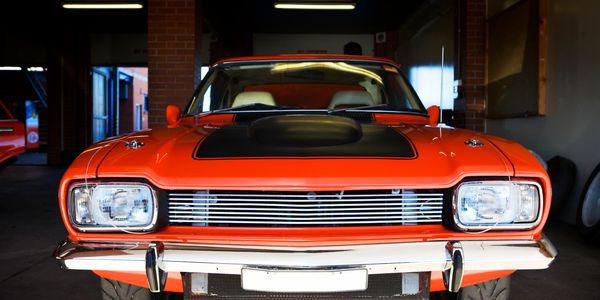 Front view of a classic orange muscle car with a black hood in a garage.