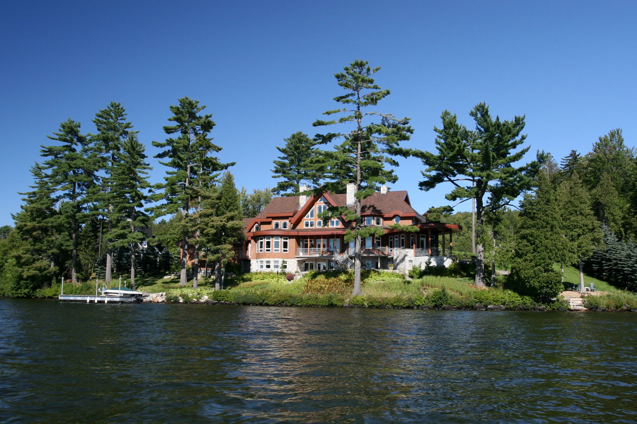Large lakeside house surrounded by tall pine trees under a clear blue sky.
