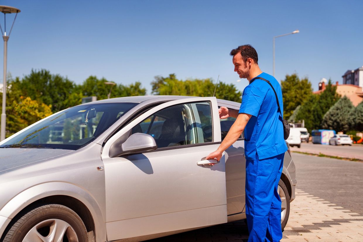 A man in blue scrubs opens a silver car door on a sunny day.