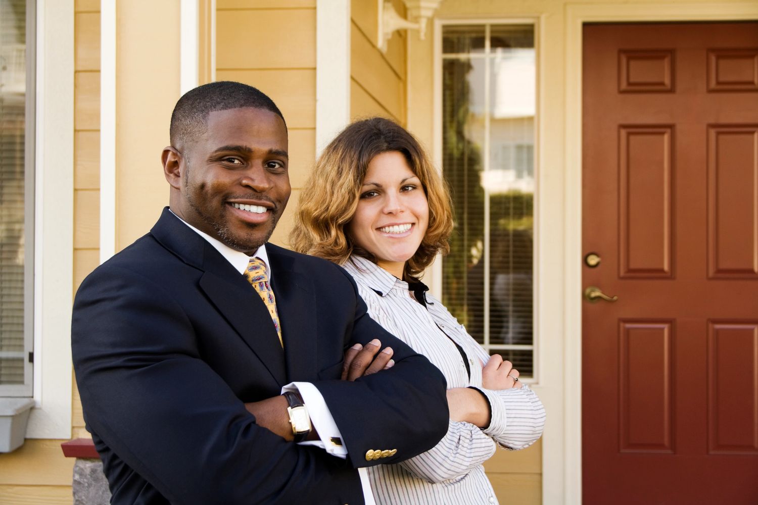 Confident man and woman standing with arms crossed in front of a house.