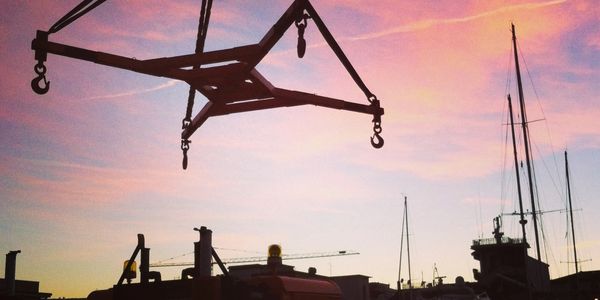 Silhouette of a crane hook against a colorful sunset sky at a harbor.