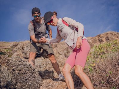 A man helps a woman climb rocky terrain during a hike under clear blue skies.