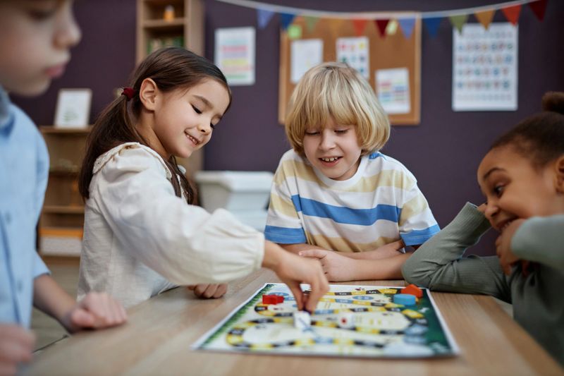 Multiethnic group of little children playing board game together in preschool and moving game pieces, copy space