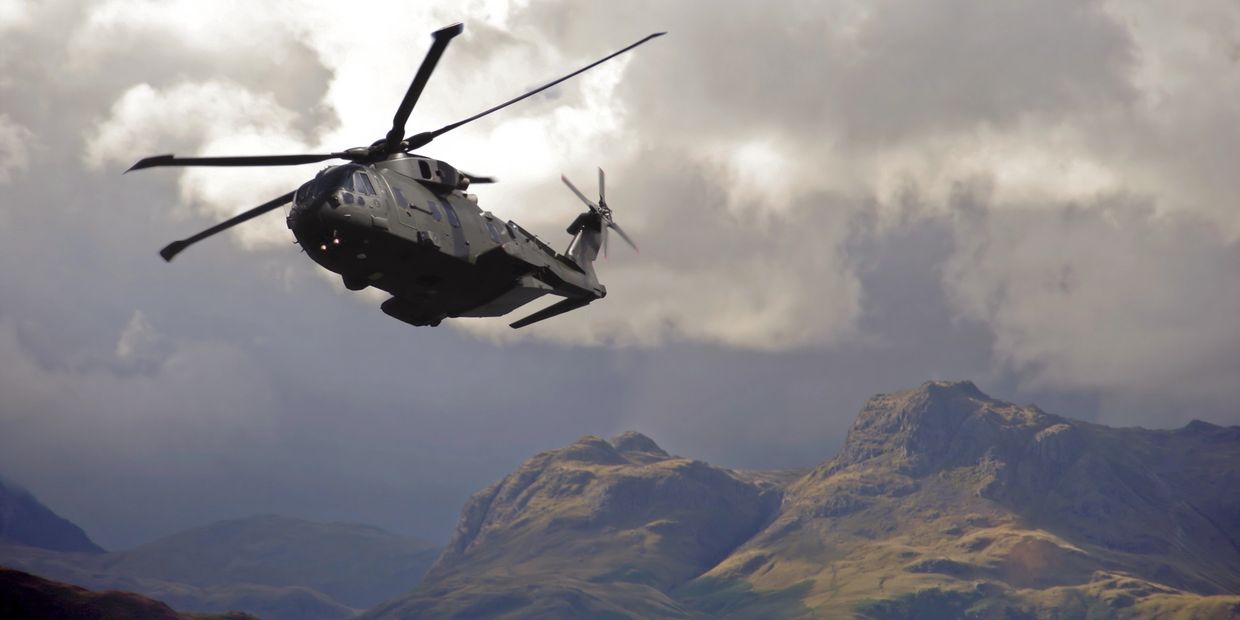Military helicopter flying over mountainous terrain under cloudy skies.