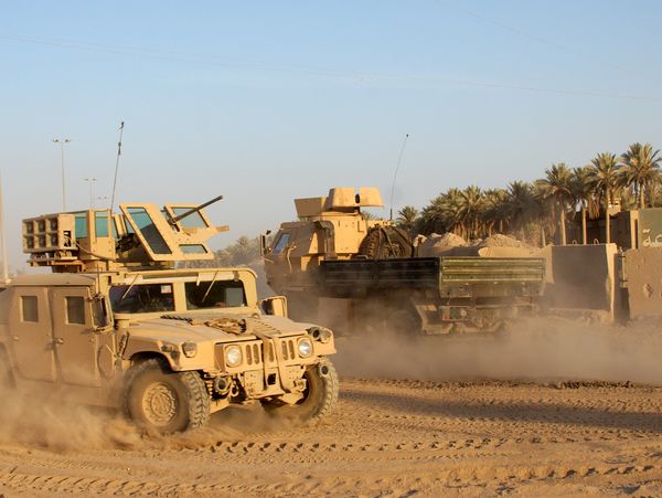 Military vehicles kicking up dust in a desert environment near a fortified area.