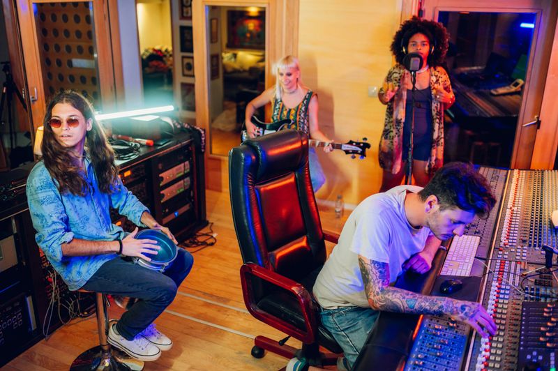 Picture of a band during a recording session in a music studio. Male percussionist with a goblet drum darbuka and a sound engineer at mixing console. Female bass player and vocalist.
