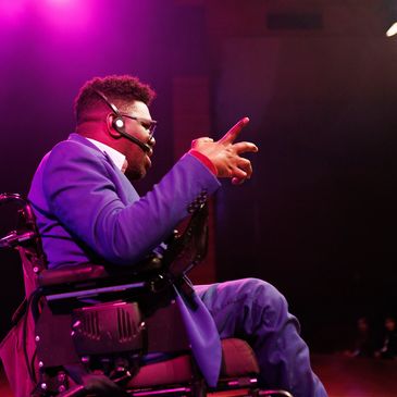 Man in wheelchair speaking on stage with headset.