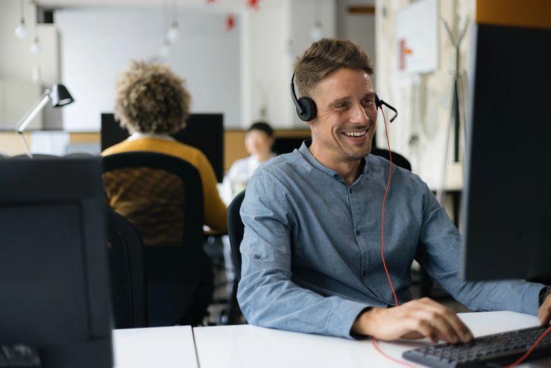 Happy man talking to someone through headset while working on desktop PC in IT support center.