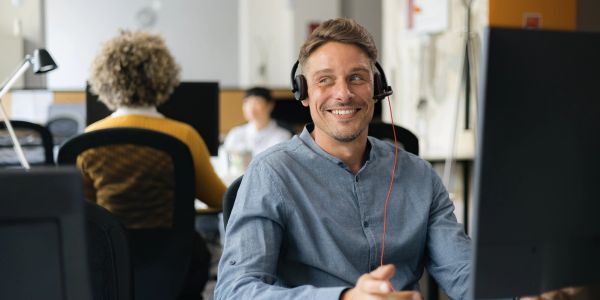 A man wearing a headset smiles at his computer in an office.