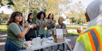 Volunteers cheerfully greet a person in safety gear outdoors.